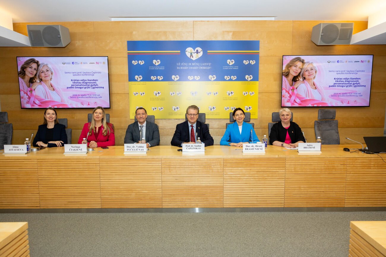 Šeši žmonės sėdi prie stalo konferencijos salėje, už jų rodomi informaciniai ekranai