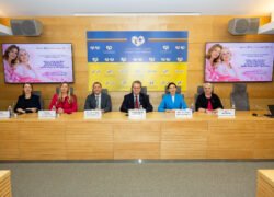 Šeši žmonės sėdi prie stalo konferencijos salėje, už jų rodomi informaciniai ekranai