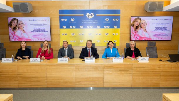 Šeši žmonės sėdi prie stalo konferencijos salėje, už jų rodomi informaciniai ekranai