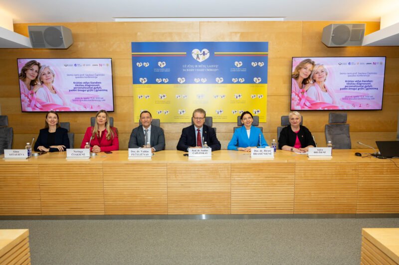 Šeši žmonės sėdi prie stalo konferencijos salėje, už jų rodomi informaciniai ekranai