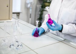 Laboratorijos darbuotojas su pirštinėmis laiko kolbą su violetiniu skysčiu