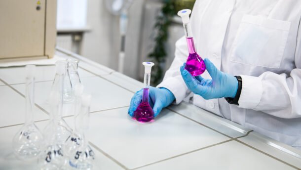 Laboratorijos darbuotojas su pirštinėmis laiko kolbą su violetiniu skysčiu