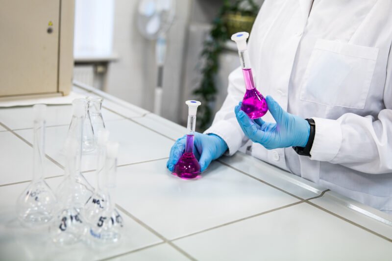Laboratorijos darbuotojas su pirštinėmis laiko kolbą su violetiniu skysčiu