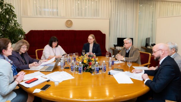 Komisijos posėdžio dalyviai sėdi prie stalo su dokumentais