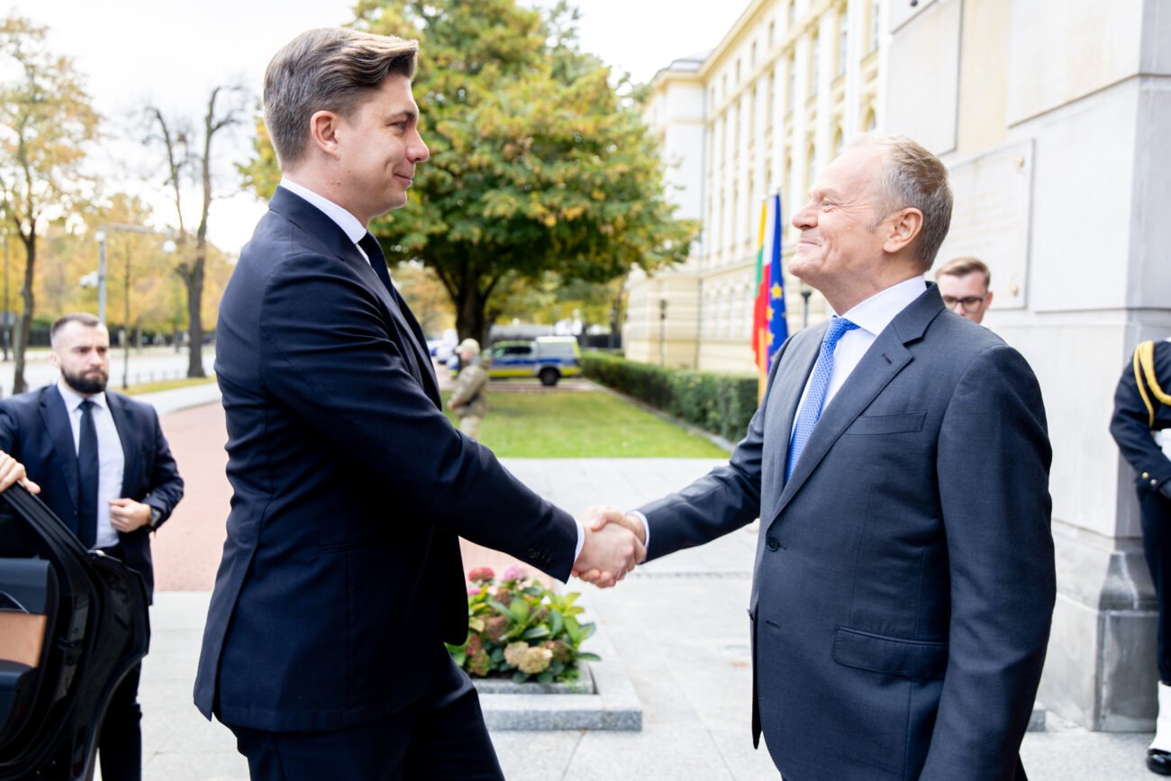 Du kostiumuoti vyrai prie pastato sveikinasi paspausdami rankas