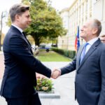 Du kostiumuoti vyrai prie pastato sveikinasi paspausdami rankas