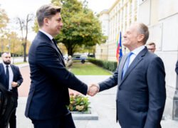 Du kostiumuoti vyrai prie pastato sveikinasi paspausdami rankas