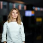 Moteris balta palaidine stovi patalpoje, fone matomas sulietas interjeras