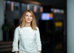 Moteris balta palaidine stovi patalpoje, fone matomas sulietas interjeras