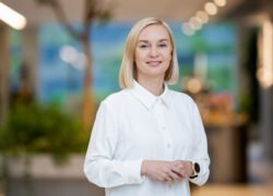 Besijuokianti šviesiaplaukė moteris baltais marškiniais stovi šviesiame interjere