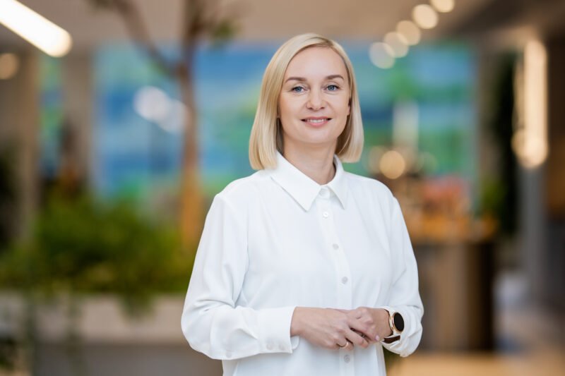 Besijuokianti šviesiaplaukė moteris baltais marškiniais stovi šviesiame interjere