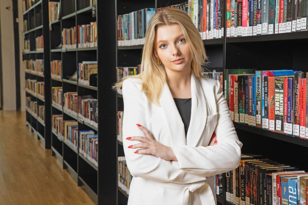 Moteris baltais drabužiais stovi bibliotekoje sukryžiuotomis rankomis