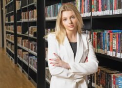 Moteris baltais drabužiais stovi bibliotekoje sukryžiuotomis rankomis