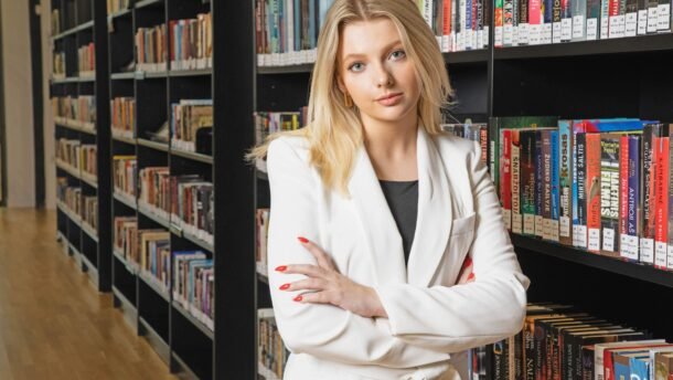 Moteris baltais drabužiais stovi bibliotekoje sukryžiuotomis rankomis