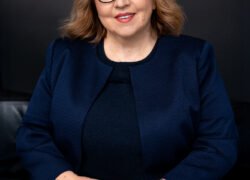 Besėdinti besišypsanti moteris su akiniais ir tamsiai mėlynu drabužiu
