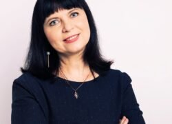 Besijuokianti moteris juodais plaukais ir tamsia palaidine sukryžiuotomis rankomis