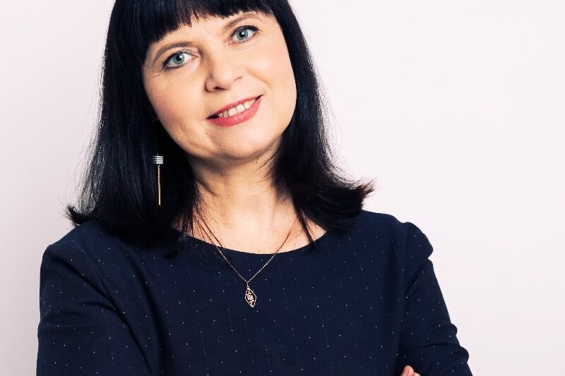 Besijuokianti moteris juodais plaukais ir tamsia palaidine sukryžiuotomis rankomis