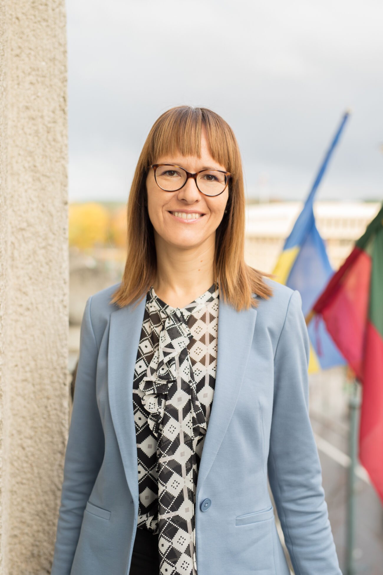 Besišypsanti moteris su akiniais stovi prie vėliavų