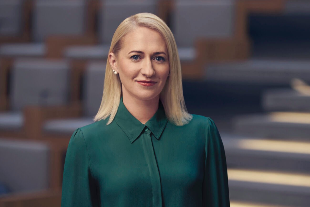 Blondinė moteris žalia palaidine stovi modernioje salėje