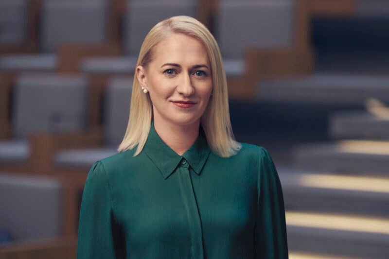 Blondinė moteris žalia palaidine stovi modernioje salėje