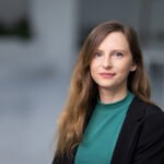 Moteris juodu švarku ir žalsvu megztiniu žvelgia į kamerą