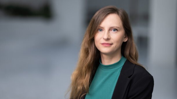 Moteris juodu švarku ir žalsvu megztiniu žvelgia į kamerą