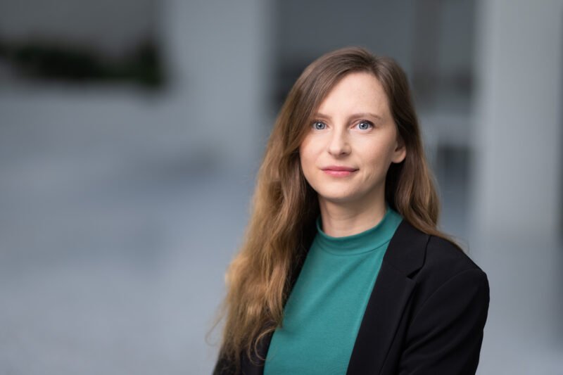 Moteris juodu švarku ir žalsvu megztiniu žvelgia į kamerą