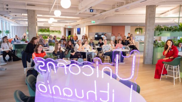 Renginio auditorija modernioje erdvėje, priekyje šviečia violetinis neoninis užrašas