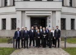 Formalioje aprangoje apsirengusių žmonių grupė stovi priešais pastato įėjimą