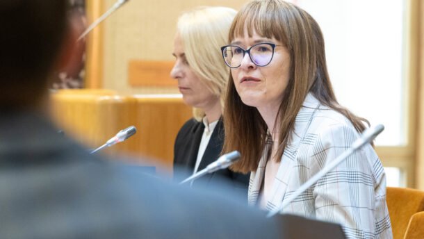 Moteris su akiniais sėdi posėdyje prie mikrofonų
