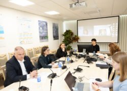 Žmonės posėdžiauja prie stalo, o ekrane vyksta vaizdo konferencija