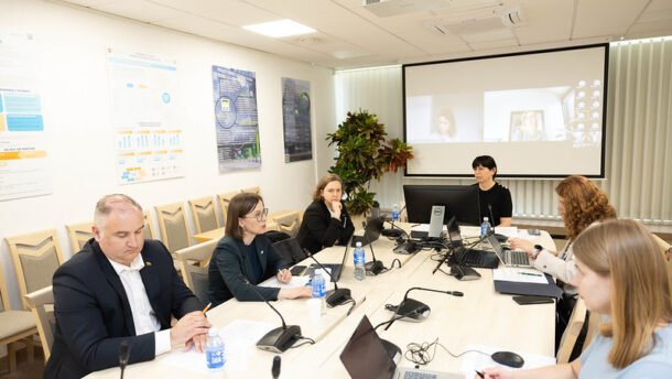 Žmonės posėdžiauja prie stalo, o ekrane vyksta vaizdo konferencija
