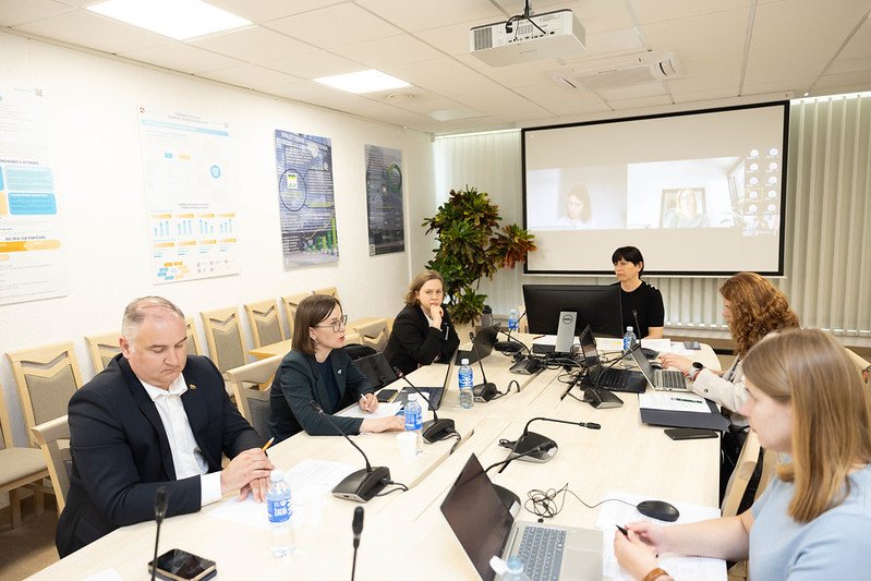 Žmonės posėdžiauja prie stalo, o ekrane vyksta vaizdo konferencija