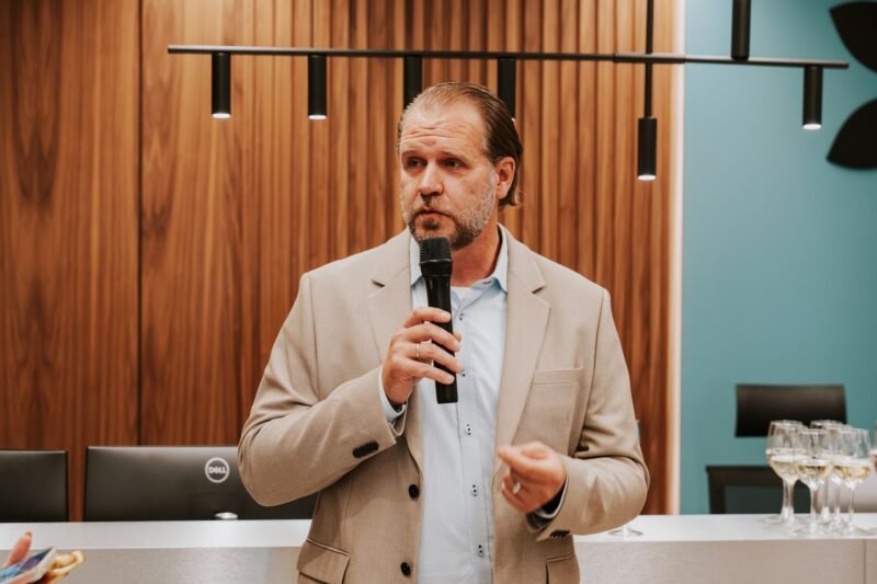 Vyras su šviesiu švarku kalba į mikrofoną modernioje renginio erdvėje