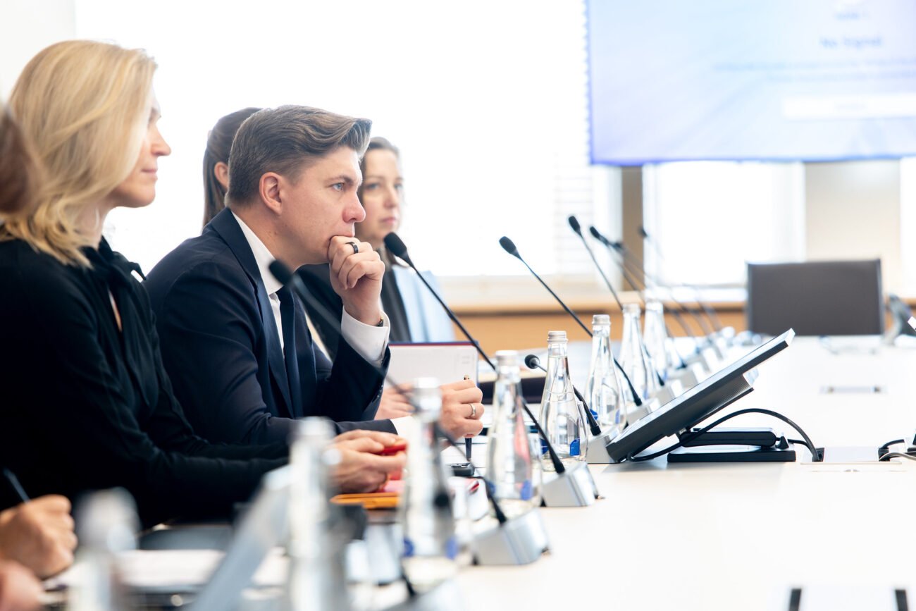 Susitikimo dalyviai sėdi prie konferencijų stalo su mikrofonais ir dokumentais