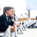 Susitikimo dalyviai sėdi prie konferencijų stalo su mikrofonais ir dokumentais
