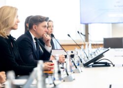 Susitikimo dalyviai sėdi prie konferencijų stalo su mikrofonais ir dokumentais