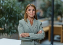 Profesionali moteris pilku kostiumu stovi biure sukryžiavusi rankas