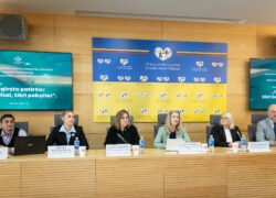 Šeši dalyviai sėdi prie konferencijų stalo su mikrofonais ir vardinėmis kortelėmis