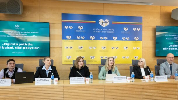 Šeši dalyviai sėdi prie konferencijų stalo su mikrofonais ir vardinėmis kortelėmis
