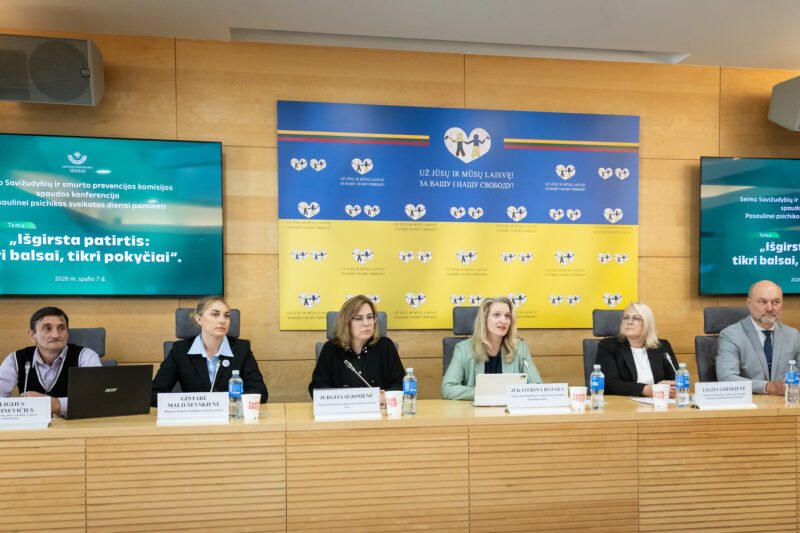 Šeši dalyviai sėdi prie konferencijų stalo su mikrofonais ir vardinėmis kortelėmis