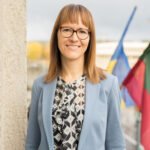 Besišypsanti moteris su akiniais ir šviesiai mėlynu švarku prie vėliavų