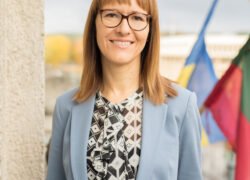 Besišypsanti moteris su akiniais ir šviesiai mėlynu švarku prie vėliavų