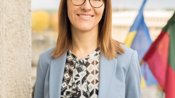 Besišypsanti moteris su akiniais ir šviesiai mėlynu švarku prie vėliavų