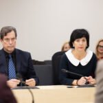 Vyras ir moteris sėdi prie posėdžių stalo bei klausosi kalbėtojo