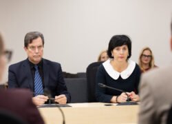 Vyras ir moteris sėdi prie posėdžių stalo bei klausosi kalbėtojo