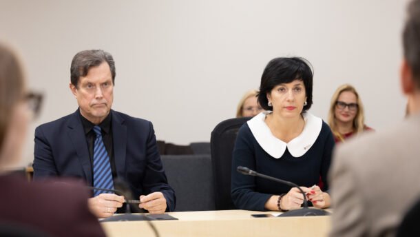 Vyras ir moteris sėdi prie posėdžių stalo bei klausosi kalbėtojo