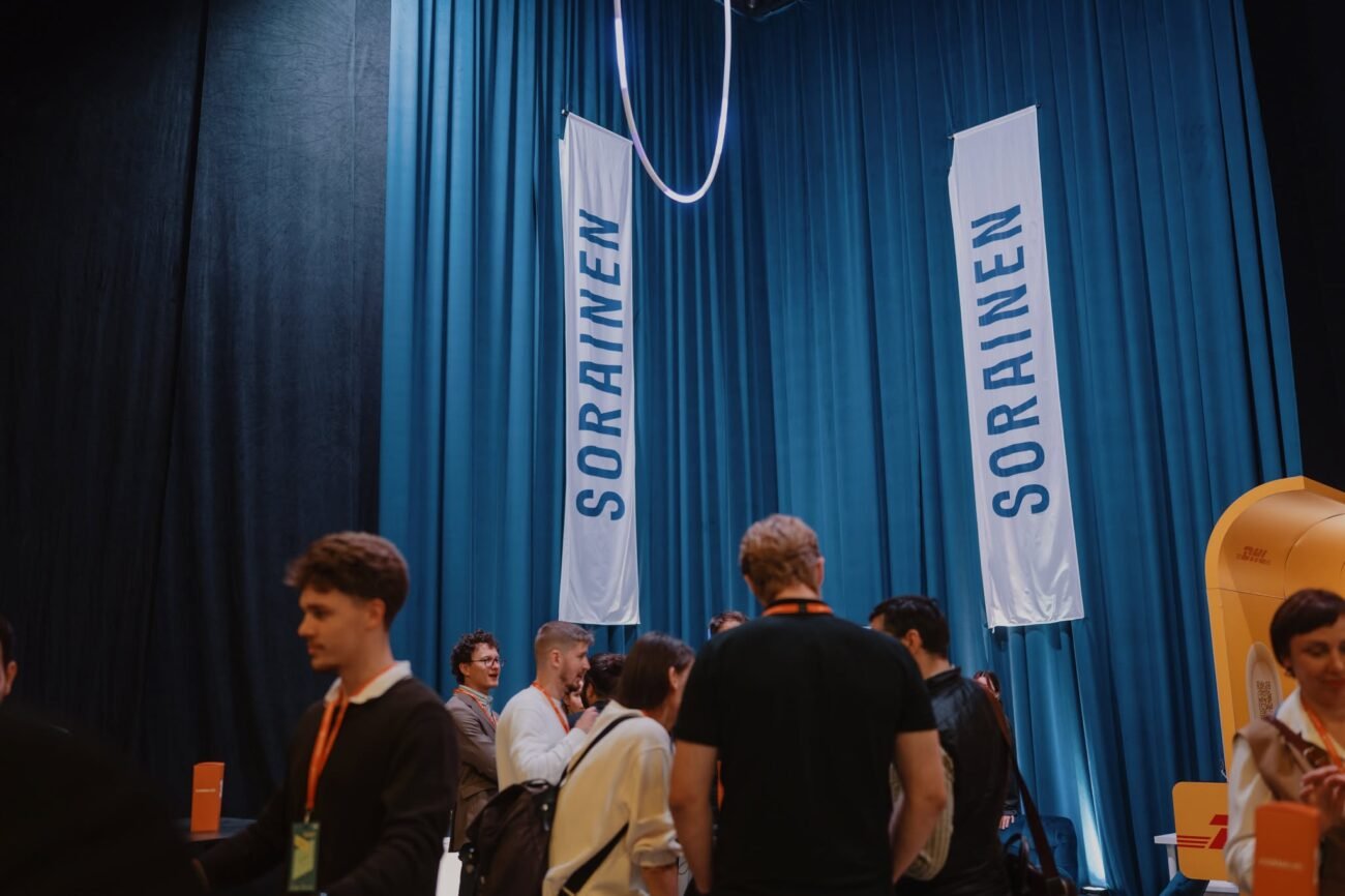 Žmonės bendrauja renginio erdvėje šalia „SORRAINE[N]“ reklaminių vėliavų