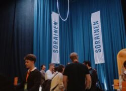 Žmonės bendrauja renginio erdvėje šalia „SORRAINE[N]“ reklaminių vėliavų