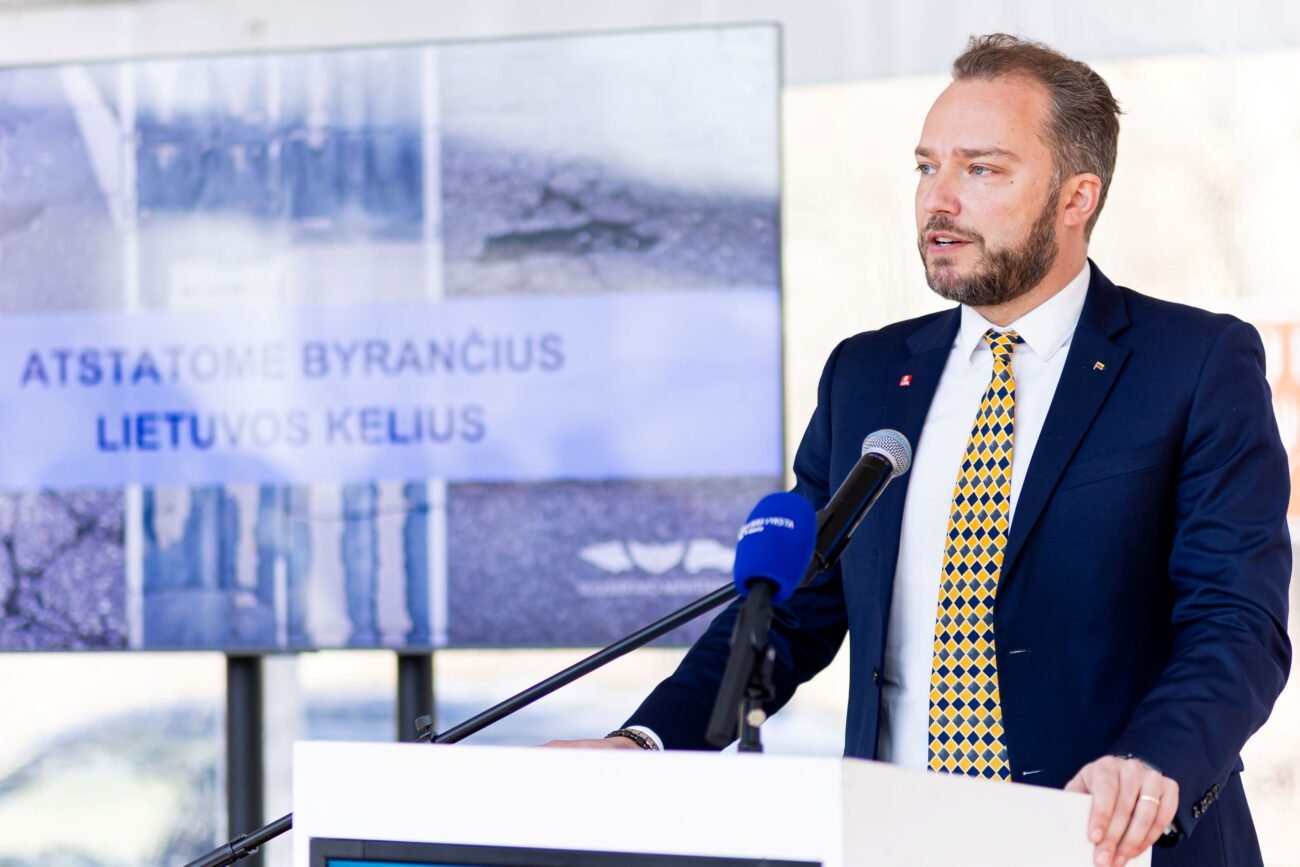 Kostiumu vilkintis vyras kalba prie tribūnos su mikrofonais